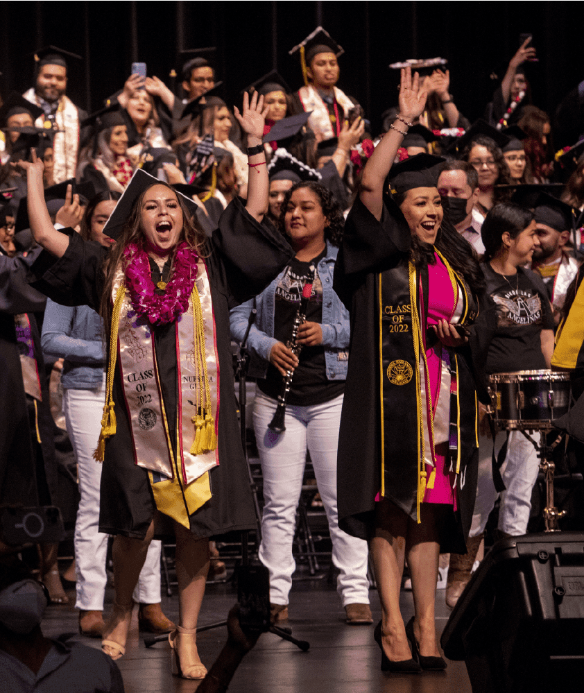 Graduates in caps and stoles cheering with raised arms onstage at a cultural graduation ceremony, a student band playing behind them