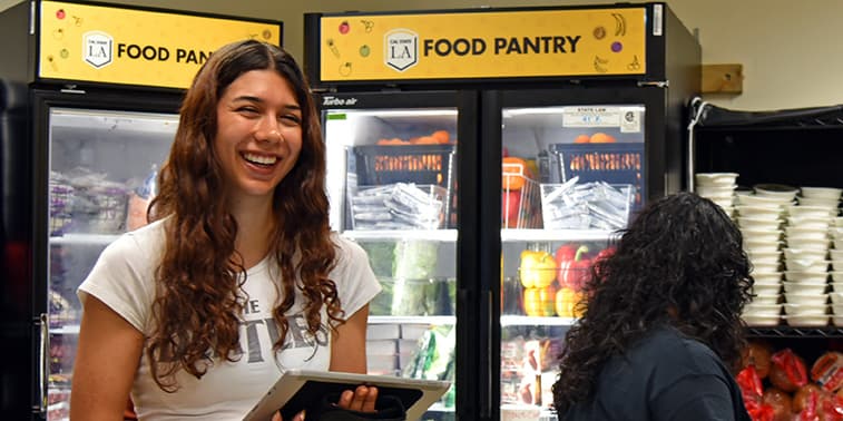 A student beside refrigerated cases stocked with produce and packaged food under a Cal State LA Food Pantry sign