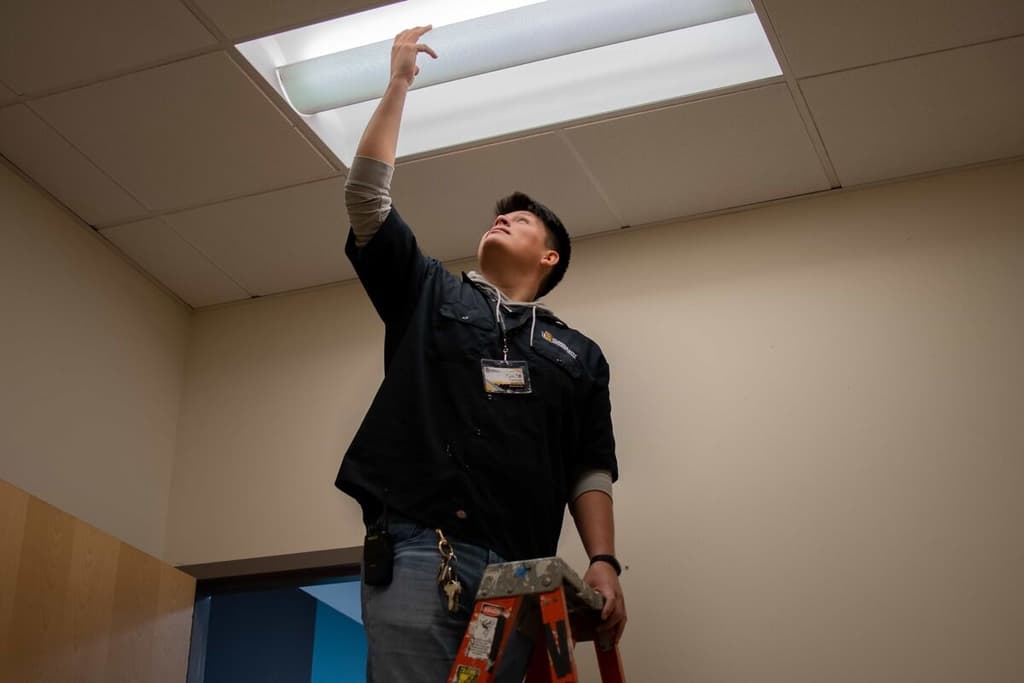 A student worker changing out a lightbulb in a ceiling fixture, standing on a ladder