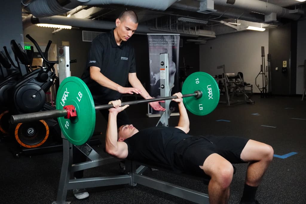 Student bench-pressing in the fitness center, with a trainer spotting them